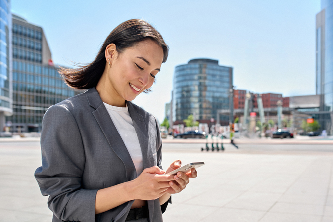 A smiling woman in a gray blazer uses her smartphone in an urban setting.